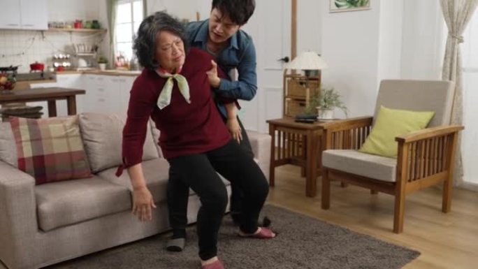 亚洲成年儿子在家里摔倒在地板上后，迅速过来帮助退休的母亲坐在沙发上休息。他在她背后放了垫子，让她感觉