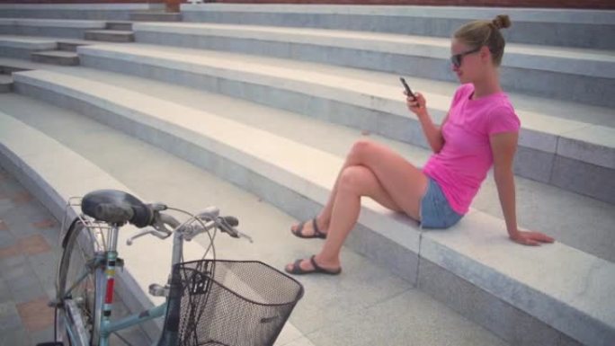 带智能手机的女孩坐在楼梯上，骑自行车日落快速慢动作