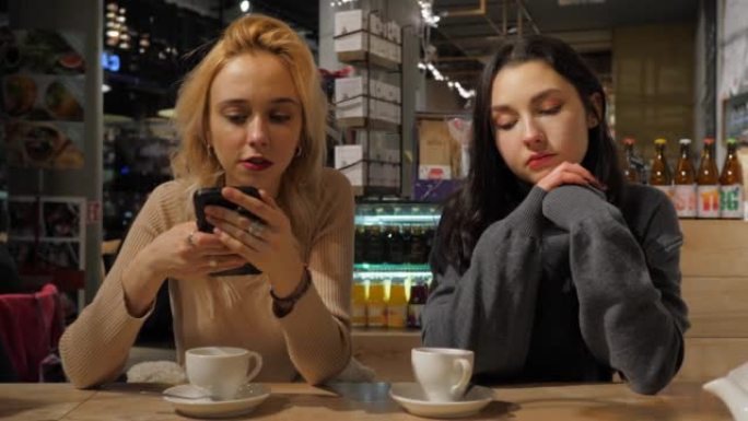 咖啡馆里的两个年轻美女使用手表智能手机聊天社交媒体，而另一个女朋友无聊喝茶