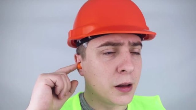 一名戴着安全帽和绿色背心的工人戴上耳塞。工业安全。工业噪声保护。在大声工作中遵守行为规则。橙色耳塞