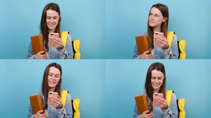 可爱的女孩青少年学生拿着书的肖像使用手机，穿着牛仔夹克和黄色背包，孤立在工作室的蓝色背景墙上。大学学