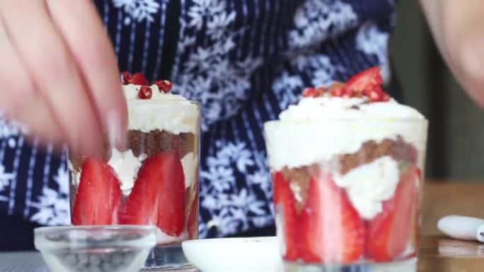 一个女人用草莓，鲜奶油和脆饼准备甜点。添加巧克力滴和冻干草莓作为装饰。特写。