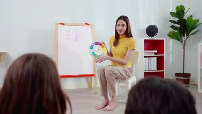 美丽的亚洲老师穿着黄色衬衫，坐在小椅子上，拿着五颜六色的时钟在教室里讲授时间课。可爱的亚洲六岁学生女