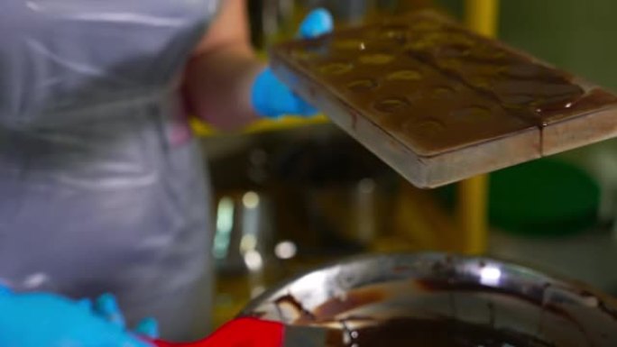 Chocolatier用最后一层巧克力覆盖填充的模具。双手戴着蓝色手套，借助刮刀从模具中取出备用巧克