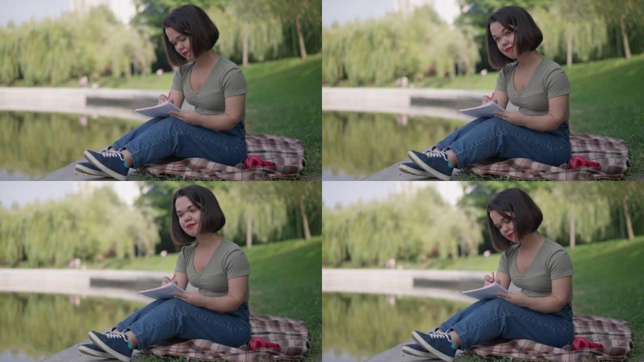 坐在公园湖岸上微笑的小女人的侧视图用笔写日记。快乐放松的高加索美女与侏儒症梦想在户外休息。