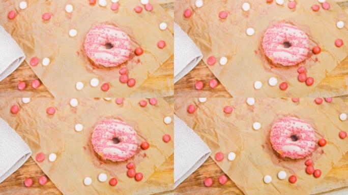 粉色甜甜圈装饰有粉色糖衣。甜甜圈在一张用糖果装饰的纸上。微距和滑块拍摄。糖果以慢动作移动。面包店和食