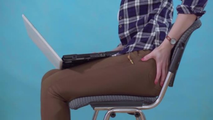 坐在蓝色背景上的女人在笔记本电脑上工作，痔疮引起疼痛和不适
