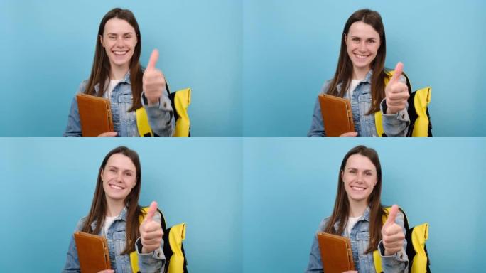 快乐女孩的肖像青少年学生穿着牛仔夹克和黄色背包拿着书显示拇指向上像手势，孤立在工作室的蓝色背景上。大