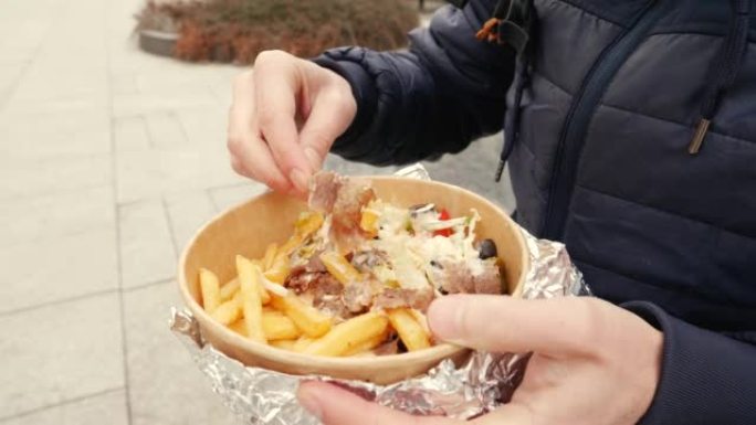 街上的男人坐在吃外卖食品盒，里面有烤肉串肉，沙拉和一次性餐具中的炸土豆片