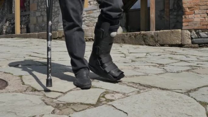 一名男子的脚踝骨折后，他的脚踝康复矫形器靠在一只拐杖上沿着道路行走。断腿用塑料紧身胸衣固定关节