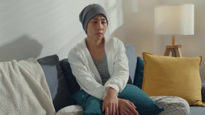 绝望的年轻亚洲癌症患者坐在家里客厅的沙发上。