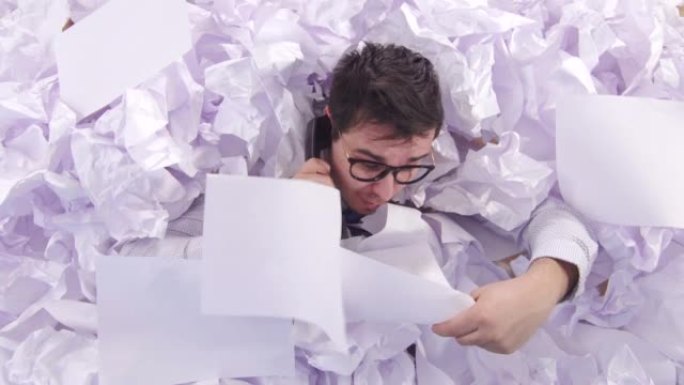 困惑的男人戴着眼镜的办公室职员正在一大堆纸上讲电话，从上面看