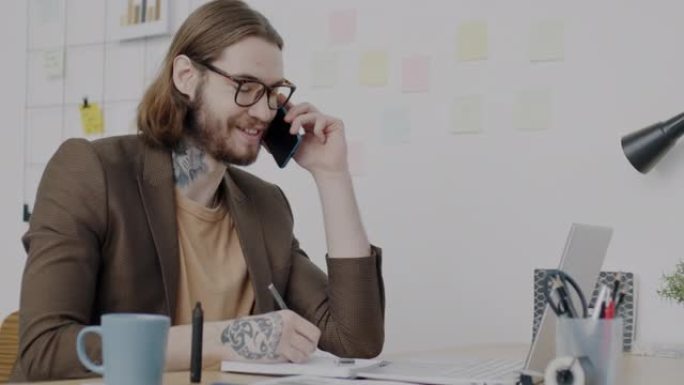 快乐的上班族在现代办公室使用笔记本电脑书写和微笑在手机上讲话