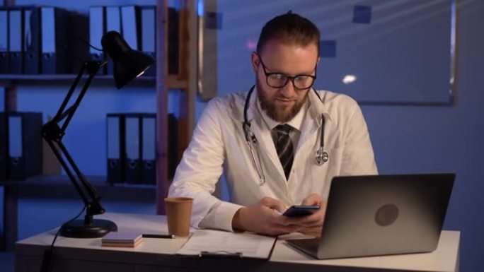 夜班医院使用移动智能手机在笔记本电脑上工作的男医生，电子健康记录系统，远程医疗