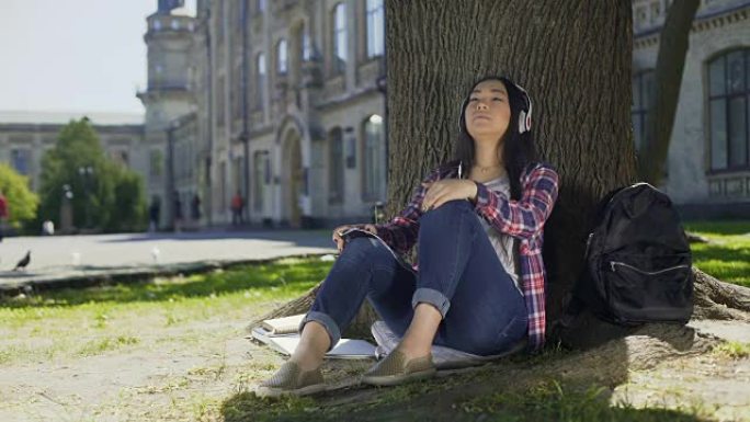 亚洲女性戴着耳机享受音乐，坐在树下，最喜欢的乐队