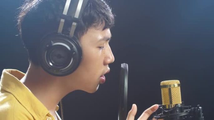 黑色背景上戴着耳机对着电容式麦克风唱歌的亚洲小男孩的特写侧视图