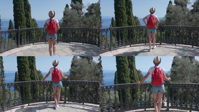戴着帽子和背包的旅游妇女在希腊科孚岛Achilleion宫的花园露台上欣赏风景，慢动作