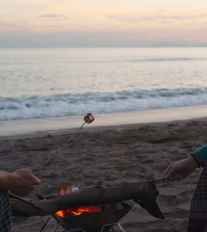 日落时分海边浪漫约会，一对年轻夫妇在篝火旁煎棉花糖。垂直视频社交媒体