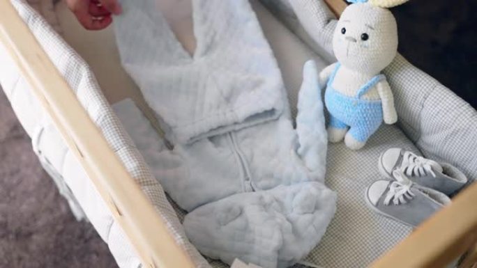 快乐的女人为男婴的到来准备必要的衣服，女性把蓝色西装放在木制婴儿床里，鞋子，针织兔子玩具和超声波图片