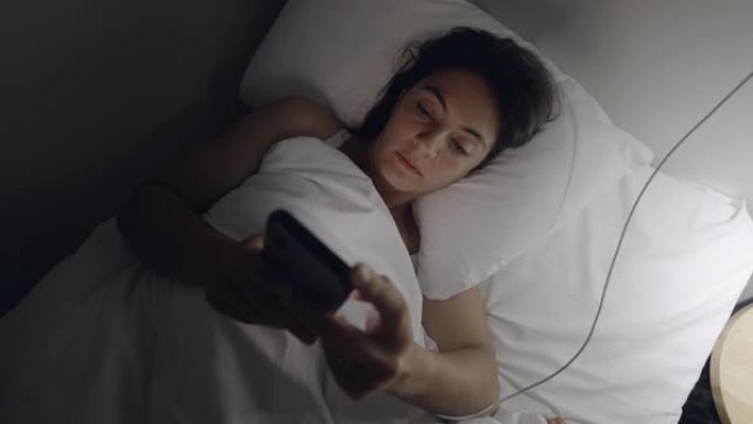 女人晚上躺在床上关掉手机。看着智能手机屏幕的女性关闭设备并关闭灯进入睡眠状态