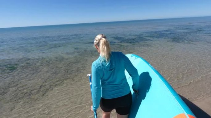 一名成熟女子背着SUP沿着海滩，走向浅滩