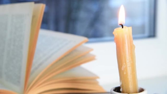 蜡烛燃烧和书在窗台上，冬天的早晨或傍晚，外面下雪，风在移动页面。选择性聚焦