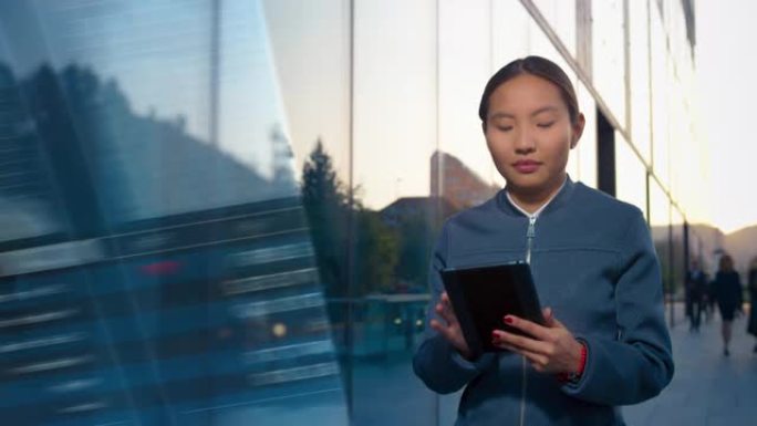 年轻的亚洲女企业家走在玻璃覆盖的商业大楼上，在她的数字平板电脑上查看股票交易应用程序