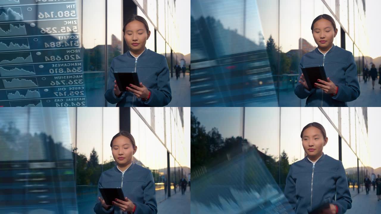 年轻的亚洲女企业家走在玻璃覆盖的商业大楼上，在她的数字平板电脑上查看股票交易应用程序