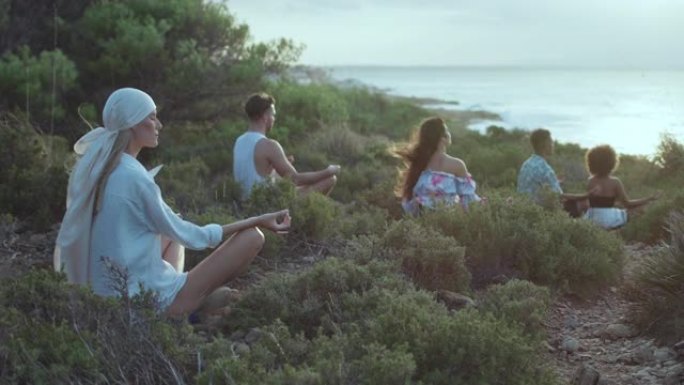 各种各样的人坐在蓝色大海旁的草崖上放松身心。莲花姿势冥想并练习健康