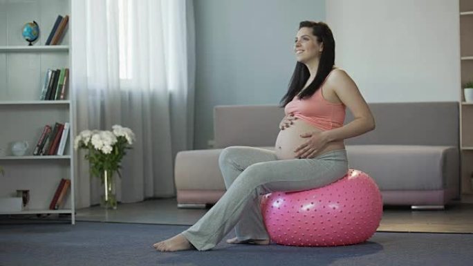 年轻的孕妇坐在健身球上，听着宝宝的跳动