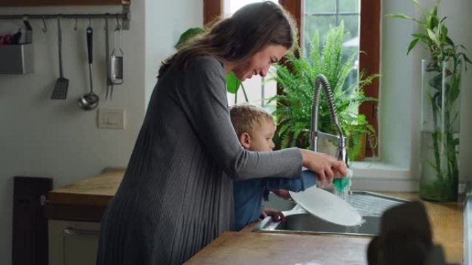 女人在教蹒跚学步的儿子如何在厨房里洗碗的手持镜头。可爱的小男孩试图帮助他的母亲做家务和任务，用海绵洗
