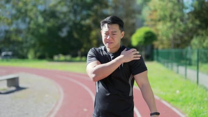 成人亚洲运动男子在城市体育场训练时感到肩膀剧痛。一个穿着运动服的悲伤男子受伤了。