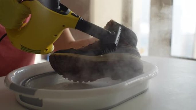 戴着橡胶手套的无法识别的男子的细节裁剪镜头使用蒸汽清洁器用热蒸汽从沙子和泥中清洗盆上肮脏的冬鞋