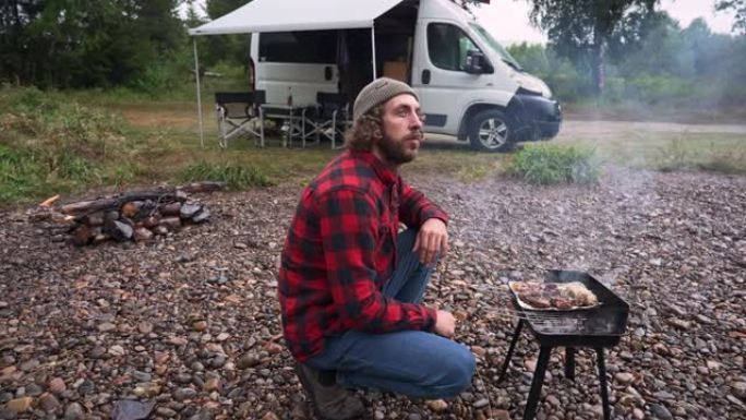 在露营车的背景下，穿着红色格子衬衫的男子在挪威河边烧烤