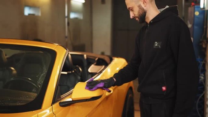 在现代洗车服务中，男性汽车服务人员的手放在黑色橡胶手套上，手握超细纤维布和清洁汽车后视镜。用超细纤维