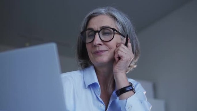 成熟的时尚公司秘书，白发和时尚的眼镜，正在考虑为客户提供正确的答案，在笔记本电脑上保持在线业务往来，
