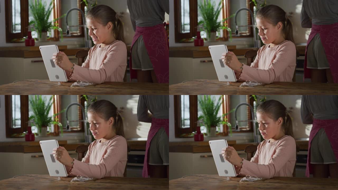 一个可爱的小女孩在厨房里使用数字平板电脑的肖像，而母亲在背景中做饭。玩教育游戏和使用技术的女孩子