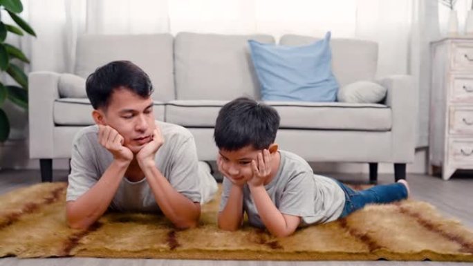 4K，一个亚洲父子在房子中间的客厅里放松，双手放在下巴上，在家庭度假时快乐地玩耍。