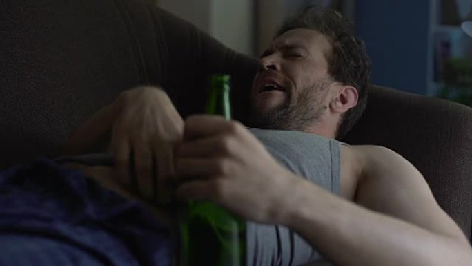 成年男子躺在沙发上，拿着啤酒瓶，打哈欠和抓挠腹部