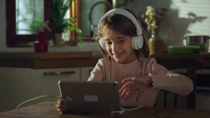 一个可爱的小女孩在厨房里用数字平板电脑和耳机与家人进行视频通话的肖像。使用技术与朋友保持远程联系的女