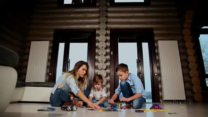 快乐的母亲和两个孩子坐在他的乡间别墅的地板上，花时间在一起收集儿童设计师。带全景窗户的客厅