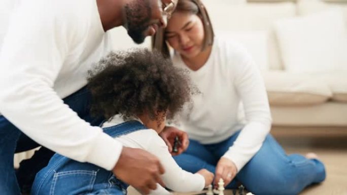 多种族的新手父母坐在地板上，和女儿在客厅里下棋。