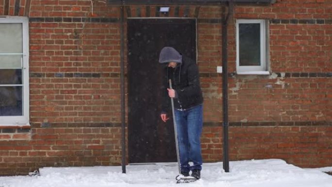 在大雪旋风慢动作中，穿着连帽衫的男子在清理房屋院子后看着破刷子