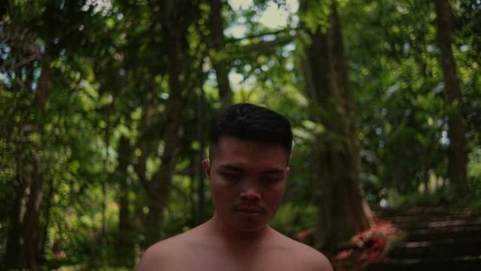 一个年轻的亚洲男子赤裸着胸部独自走在森林里，看着相机