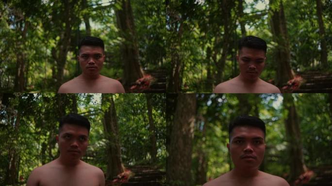 一个年轻的亚洲男子赤裸着胸部独自走在森林里，看着相机