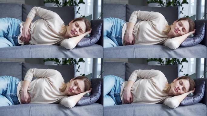 生病的年轻金发女子患有胃痛躺在沙发上抱着肚子在家里的客厅感觉胃炎或月经疼痛