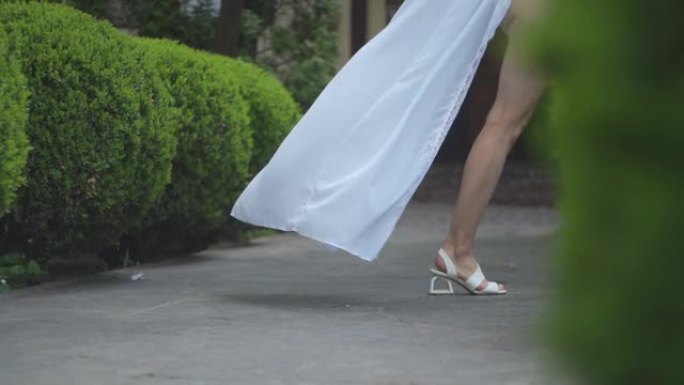穿着透明白色连衣裙和白色鞋子的女孩的腿在花园里行走。慢动作。