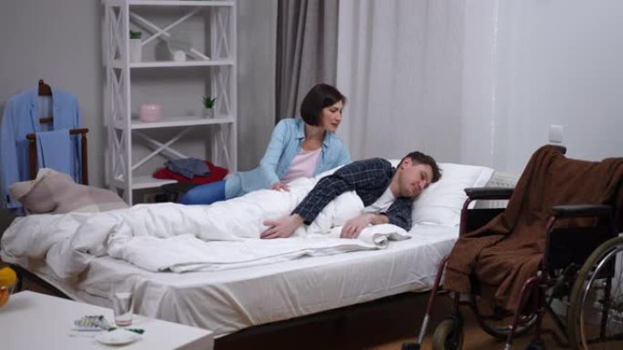 宽阔的白人男子躺在床上，作为有爱心的女人，使配偶的身体瘫痪。悲伤的丈夫和体贴的妻子在家里的卧室里坐着