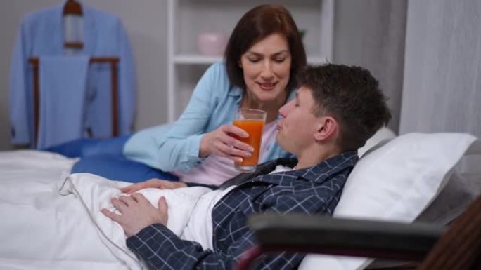 侧视图白人男子躺在床上喝橙汁，女子帮忙说话。微笑的爱的妻子在家里的卧室里照顾生病的丈夫。