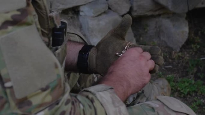 一名军人身穿迷彩服，戴着战术手套，手里拿着徽章和金属链上的十字架，在战斗前把它压在胸前，压在心上祈祷